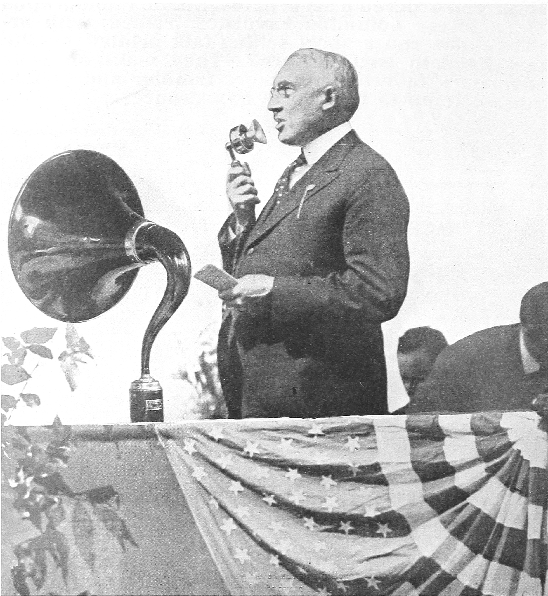A speaker at a flag-draped outdoor podium using an early microphone beside a Magnavox horn loudspeaker