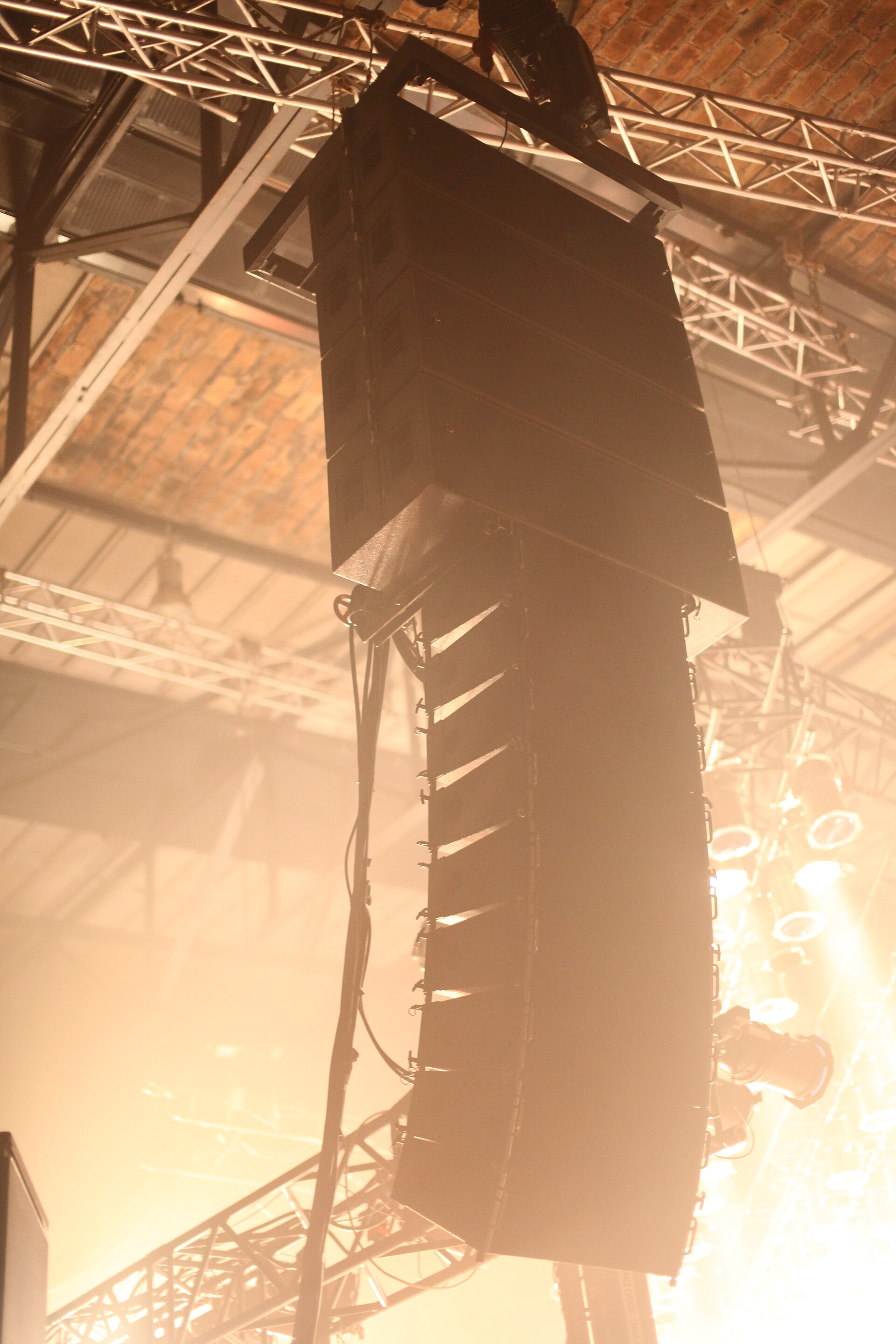 A flown line-array loudspeaker cluster suspended from overhead truss in stage lighting and haze
