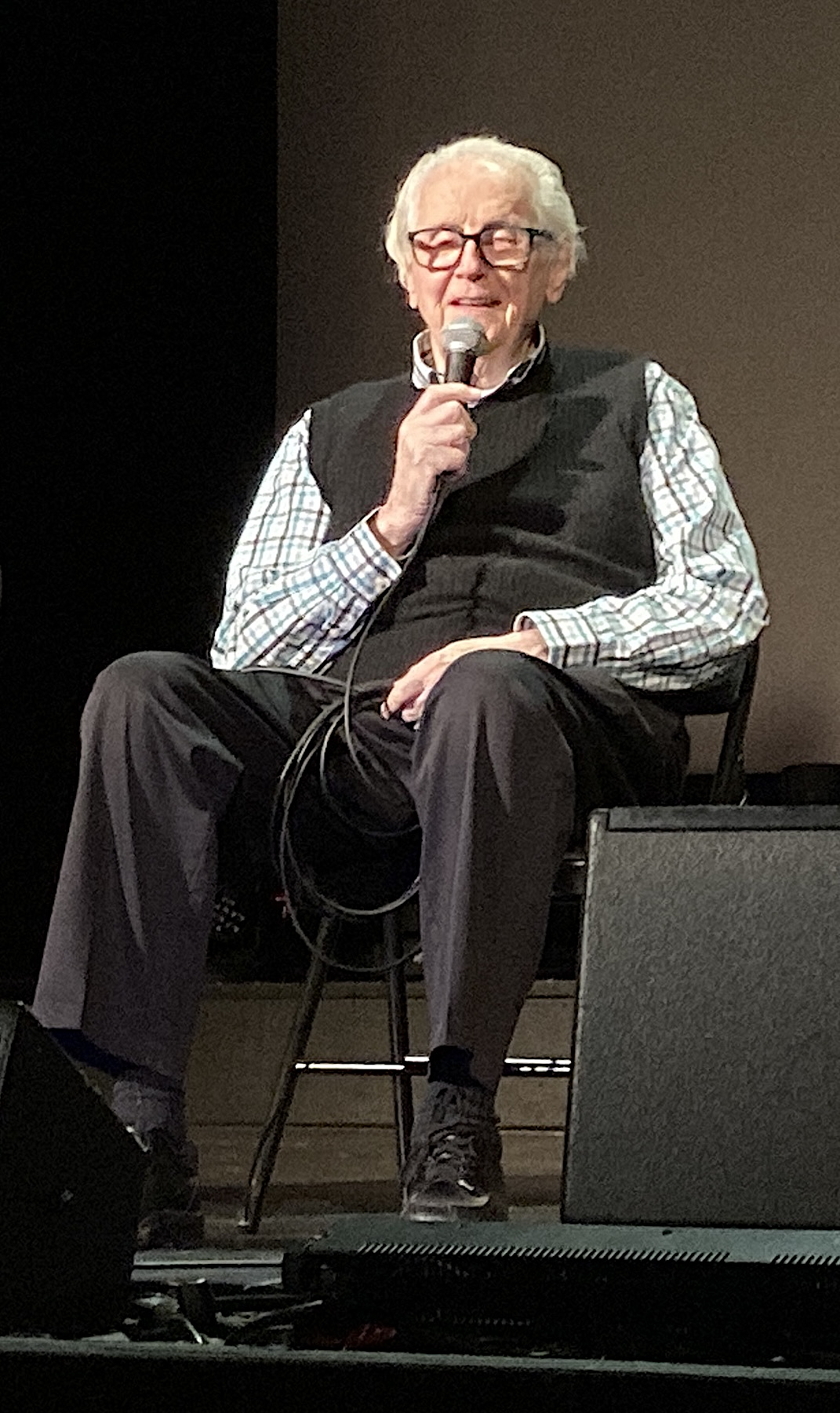 An elderly sound engineer seated on a stage speaking into a handheld microphone with wedges in front