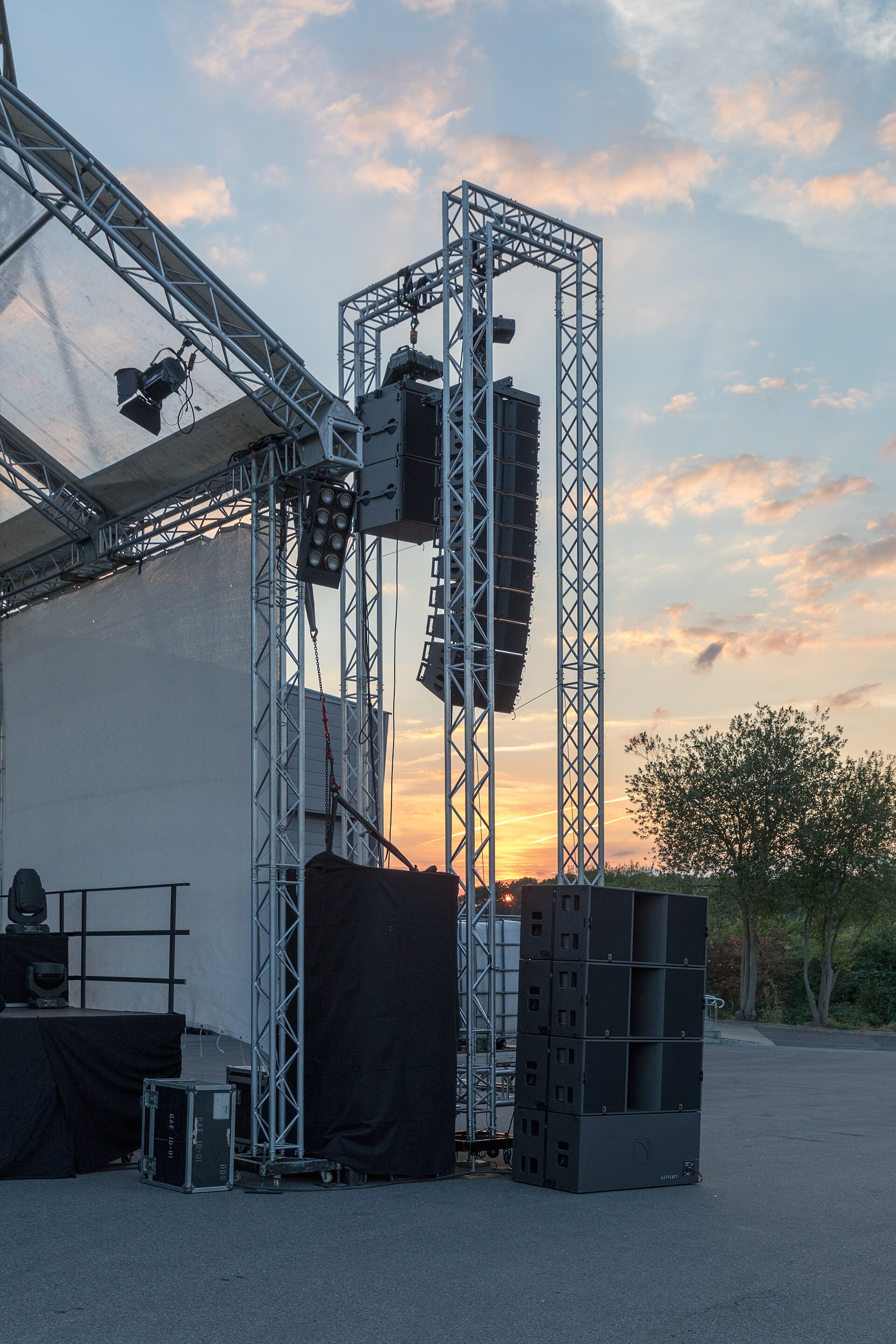 An outdoor festival stage at dusk with flown arrays, a subwoofer stack and road cases